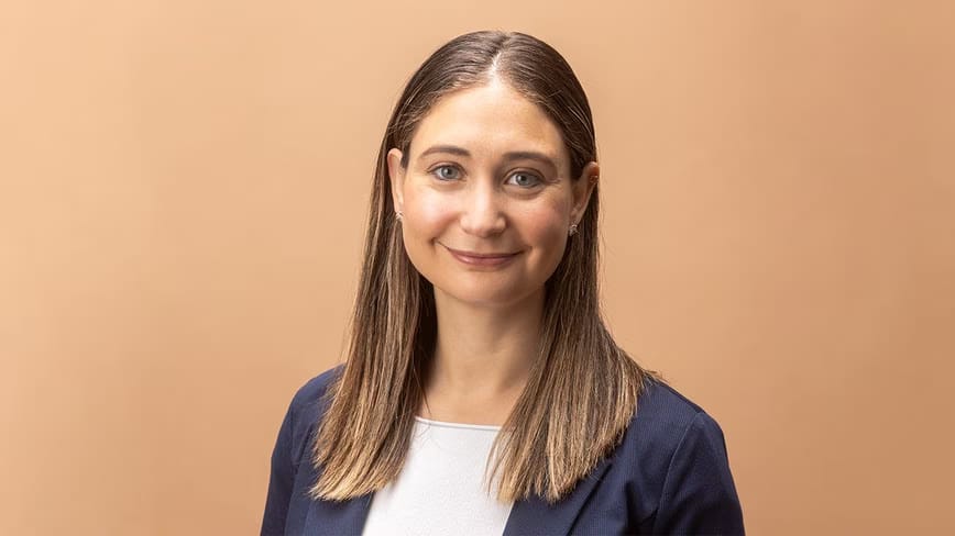 Daniela Amodei, with long hair and a navy blazer over a white top, smiling against a peach background