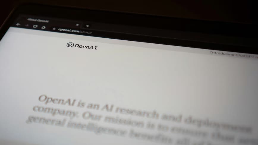 Close-up of a laptop screen showing the OpenAI logo and the openai.com about page, its mission statement blurred in the foreground