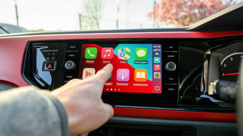 A finger pointing at the Home Screen, showing apps like Maps and Podcasts in Apple CarPlay.
