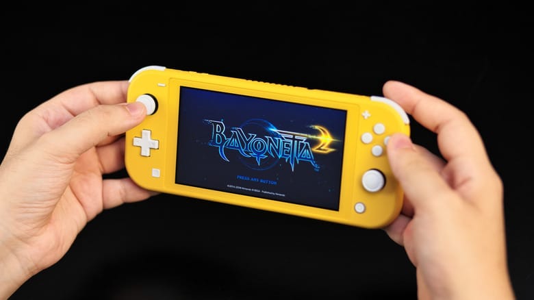 Hands holding a yellow Nintendo Switch Lite, showing the Bayonetta 2 title screen.